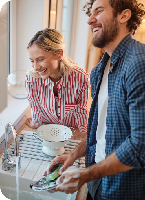 A young couple laughs while washing dishes together.